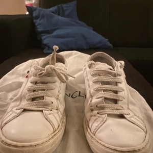 Vita sneakers från Moncler - Snygga vita sneakers från Moncler med klassisk snörning och logotypdetalj. Perfekta för en stilren look. Skick 7/10 då bakre delen på höger sko är trasig, dustbag till skora kommer med! 1000kr kommer andvändas som en startgrund för pris, dock kan pris diskuteras privat