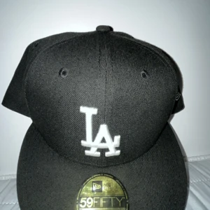 Svart keps från New Era - Snygg svart keps från New Era med LA-logga i vitt framtill. Perfekt för en casual look eller sportiga tillfällen. Kepsen har en klassisk design med platt skärm och broderade ventilationshål.