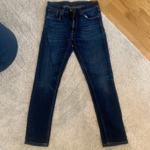 Mörkblå jeans från Nudie Jeans - Säljer ett par mörkblå jeans från Nudie Jeans med klassisk femficksdesign och orange sömmar. Jeansen har en slim passform och är tillverkade i denim. Perfekta för en stilren look!