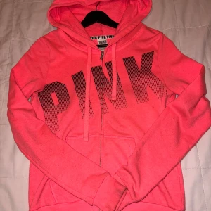 Rosa hoodie från Victoria's Secret PINK - Säljer en snygg rosa hoodie från Victoria's Secret PINK med dragkedja och stor text på framsidan. Den är tillverkad i en mjuk bomulls- och polyesterblandning och har en bekväm huva. Perfekt för en avslappnad stil!
