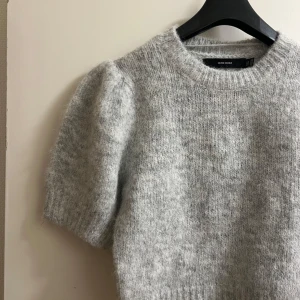 Grå stickad tröja från Vero Moda - Mysig grå stickad tröja från Vero Moda med korta ärmar och rund hals. Perfekt för kyliga dagar när du vill ha något bekvämt och stilrent. Tröjan har en mjuk och fluffig textur som ger en extra touch av komfort.