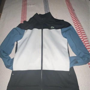 Svart och vit jacka från Lacoste - Snygg sportig jacka från Lacoste i svart och vit med blå ärmar. Jackan har en dragkedja framtill och det klassiska Lacoste-logotypen på bröstet. Perfekt för en casual look.