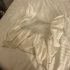 Vit blus från Zara  - Söt vit blus från Zara med smockdetaljer och volang. Perfekt för sommarens varma dagar. Klänningen har en luftig passform och är ärmlös, vilket gör den bekväm och stilren. För liten på mig så det kanske ser lite konstigt ut på bilden.