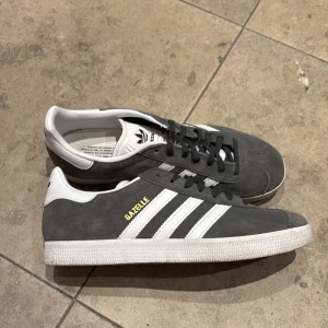 Grå Adidas Gazelle sneakers - Snygga grå Adidas Gazelle sneakers med vita detaljer och klassiska tre ränder. Skorna har en stilren design med snörning och en bekväm passform. Perfekta för en avslappnad stil.