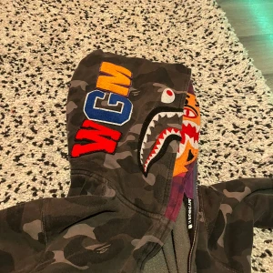 Camouflage hoodie med hajmotiv - Säljer en cool camouflage hoodie från A Bathing Ape med ett hajmotiv på huvan. 💕Hoodien har en dragkedja framtill och färgglada detaljer i lila och orange. Perfekt för dig som vill sticka ut! Den är bra skick storlek L bara fråga om fler bilder eller frågor så svarar jag💕köpte den från min kusin först för 900 eller vad det va💕