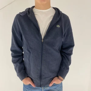 Mörkblå hoodie från Lacoste Sport - Säljer en mörkblå hoodie från Lacoste Sport med dragkedja och huva. Den har det klassiska Lacoste-märket på bröstet. Passar som en M/L. 