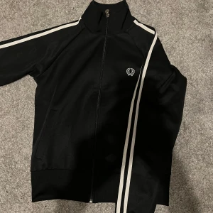 Svart track jacket från Fred Perry - Snygg svart track jacket från Fred Perry med vita ränder längs ärmarna och broderad logga på bröstet. Jackan har dragkedja och ribbade muddar. Perfekt för en casual stil. Storleken är 38 vilket skulle motsvara 150-160 centimeter. Tveka inte på att höra av er vid några som helst funderingar.