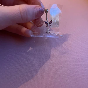 Snygg navelpiercing med lila och klara stenar - Säljer en elegant navelpiercing med en böjd stav och en kula i silver. Smycket har två lila stenar formade som en fjäril och en klar, gnistrande blomma som hänger nedanför. Perfekt för att ge en extra touch till din stil.