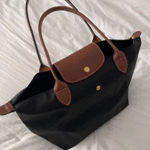Longchamp Svart Le Pliage Original M Tote Bag - Säljer en stilren svart Le Pliage Original M Tote Bag från Longchamp. Väskan är tillverkad i återvunnen canvas med bruna skinnhandtag och en praktisk dragkedja. Perfekt för att bära med sig allt man behöver under dagen.