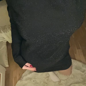 Svart glittrig klänning med öppen rygg - Snygg svart glittrig klänning med och en öppen rygg med knytning. Klänningen har långa ärmar och en tight passform som framhäver figuren.