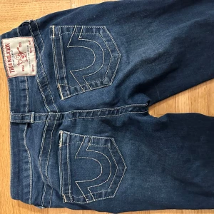 Blå jeans från True Religion - Säljer ett par blå jeans från true religion. Jeansen har de klassiska bakfickorna och är lågmidjade o bootcut. Tyget precis vid gylfen är trasigt men går nog lätt att sy för hand. 