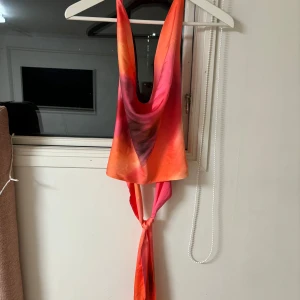 Färgglad topp från BikBok - Säljer en färgglad topp från BikBok i storlek M. Toppen har ett livfullt mönster i orange, rosa och lila nyanser. Den är ärmlös och har en knytning i ryggen för en snygg detalj.