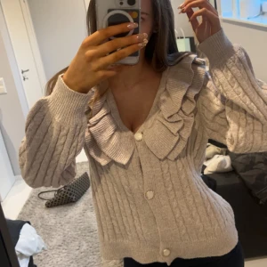 Beige stickad kofta med volangdetaljer - Säljer en mysig beige stickad kofta med volangdetaljer längs kragen. Koftan har långa ärmar och knäpps med knappar framtill. Perfekt för kyliga dagar!