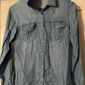 Blå jeansskjorta från Vila - Snygg blå jeansskjorta från Vila med klassisk krage och knappar framtill. Skjortan har två bröstfickor med knappstängning och långa ärmar. Perfekt för en avslappnad stil.