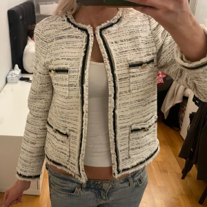 Vit kavaj/kofta - Snygg vit kavaj med svarta detaljer och guldknappar. Den har en klassisk design med fyra fickor framtill och en rund halsringning. Perfekt för en stilren look. Några av spännerna är trasiga men annars är den i bra skick, säljer därför billigt