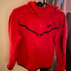 Röd zip hoodie från Nike tech junior stl. L - Snygg röd jacka från Nike med svarta detaljer och dragkedja. Perfekt för en sportig look. Jackan har långa ärmar och en bekväm passform. Storlek L motsvarar 147-158