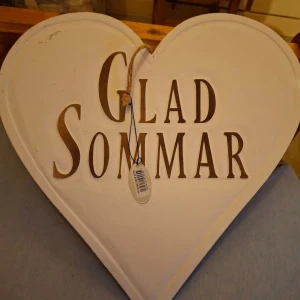 Vit hjärtformad skylt med texten 'Glad Sommar' - Charmig vit hjärtformad skylt med texten 'Glad Sommar' i brunt. Perfekt för att hänga upp som dekoration under sommarmånaderna. Skylten är 35 cm stor och har ett snöre för enkel upphängning.