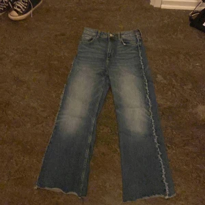 Blå jeans med fransade kanter - Säljer ett par blå jeans med fransade kanter och en unik detalj längs sidorna. Byxorna har en bootcut-stil och är perfekta för en avslappnad look. De har klassisk femficksdesign och en knappgylf.
