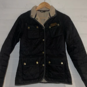 Svart quiltad jacka från Barbour - Snygg svart quiltad jacka från Barbour med beige foder. Jackan har flera fickor med knappar och en klassisk design. Perfekt för kyligare dagar.