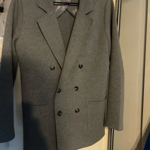 Grå kavaj från John Henric - Snygg grå kavaj från John Henric i klassisk design med dubbelknäppning. Perfekt för en stilren look. Kavajen har långa ärmar och är tillverkad i ett mjukt material. Tveka inte på att höra av dig!