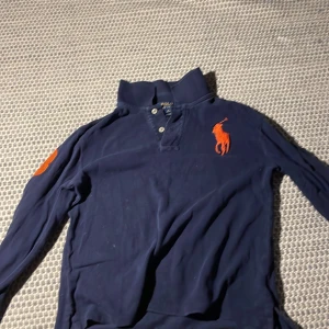Mörkblå pikétröja från Ralph Lauren - Säljer en snygg mörkblå pikétröja från Ralph Lauren med orange logga och nummerdetalj på ärmen. Tröjan har knappar vid halsen och långa ärmar. Perfekt för en stilren look!