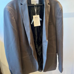 Ny kavaj från By TeeShoppen - Snygg brun kavaj från By TeeShoppen i modellen PKTHKT Phil Jersey Blazer. Perfekt för en stilren look med klassiska detaljer som knappar och fickor. Passar utmärkt till både formella och avslappnade tillfällen. Aldrig använd och har prislapp kvar. Nypris: 600kr