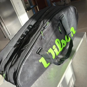 Svart Wilson tennisväska - Säljer en svart Wilson tennisväska med gröna detaljer och dragkedjor. Väskan har flera fack för att organisera utrustning och är perfekt för att bära tennisracketar och tillbehör. Den har bekväma handtag för enkel transport. 4-6 tennisrack. Nypris ca 1300kr