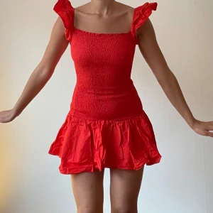 Röd sommarklänning med volangdetaljer - Säljer en superfin röd klänning perfekt för sommaren! Den har en tight passform upptill med smock och volangdetaljer på axlarna. Kjolen är kort och har en härlig volangkant. Perfekt för både fest och vardag. 🥰