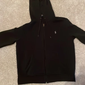 Svart hoodie från Ralph Lauren - Säljer min Ralph lauren full zip. Väldigt snygg färg och matchar med mycket. Skicket är 9/10. Väldigt skön att ha på sig och är bra för hösten och vintern. Du kan också ha den hemma som en chill tröja.