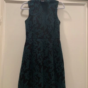 Svart och grön ärmlös klänning - Snygg ärmlös klänning i svart och grön med ett vackert blommigt mönster. Den har en dragkedja baktill och är perfekt för både fest och vardag. Klänningen är knälång och har en figurnära passform som framhäver midjan.