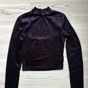 Svart glittrig topp med öppen rygg - Snygg svart glittrig långärmad topp med lila glitter och en öppen rygg. Perfekt för fest tillfällen! Materialet är mjukt och stretchigt, vilket gör den bekväm att bära hela kvällen. Toppen har en hög krage som käpps där bak. Skriv gärna om du är intresserad innan du köper 🫶