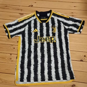 Juventus fotbollströja med Vlahović 9 - Säljer en Juventus fotbollströja i nyskick med Vlahović nummer 9 på ryggen. Tröjan är kortärmad och har ett coolt svartvitt randigt mönster med gula detaljer. Tillverkad av Adidas med Jeep-logga på framsidan. Perfekt för alla Juve-fans! ⚽️