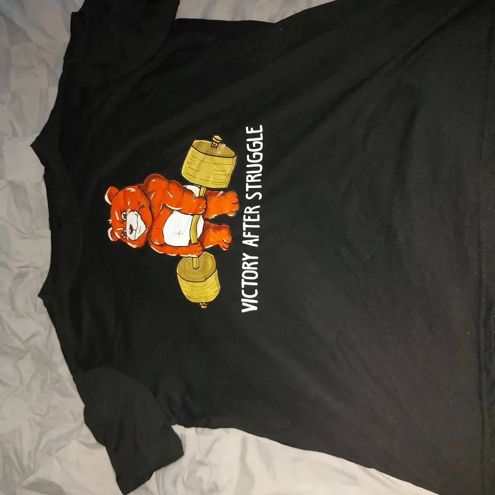 Säljer en svart t-shirt från Rejune i storlek XL. Den har ett coolt tryck med en orange björn och texten 'Victory After Struggle'. Perfekt för en avslappnad stil eller pump cover på gymmet🏋‍♂️. T-shirten är i bra skick och gjord av mjukt material.. T-shirts.