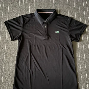 Svart pikétröja från Lacoste - Säljer en klassisk svart pikétröja från Lacoste i mycket bra skick. Tröjan har korta ärmar och den ikoniska krokodilloggan på bröstet. Perfekt för både vardag och lite finare tillfällen. Passar till allt! 🐊