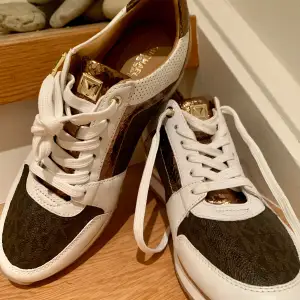 Snygga Michael Kors sneakers i vitt och guld med bruna detaljer. Skorna har en glittrig finish och snörning framtill. Perfekta för att ge din outfit en lyxig touch. De har en bekväm passform och är i mycket bra skick. Perfekta för både vardag och fest!