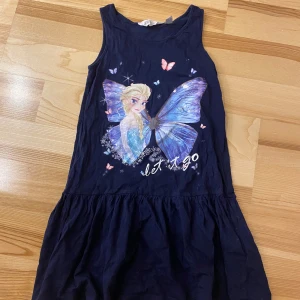Mörkblå klänning med Elsa-tryck från H&M - Söt mörkblå klänning från H&M med ett stort Elsa-tryck från Disney's Frozen. Klänningen är ärmlös och har en avslappnad passform, perfekt för sommarens äventyr. Den är dekorerad med fjärilar och texten 'let it go'.