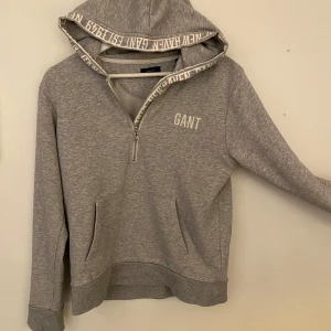 Grå hoodie från GANT - Säljer en snygg grå hoodie från GANT i storlek 170 cm. Den har en halv dragkedja och en bekväm huva med textdetaljer. Perfekt för både vardag och chill. Tröjan är i bra skick. Ny pris 999‼️