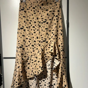 Beige kjol med svart mönster - Snygg beige kjol med svart prickigt mönster och volangdetaljer. Kjolen har en omlott-design som ger en fin rörelse och stil. Perfekt för att ge en trendig touch till din outfit.