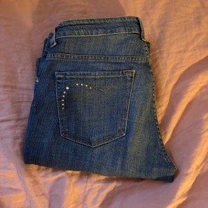 Blå jeans med bootcut - Snygga blå jeans med bootcut-stil och dekorativa stenar på bakfickorna. Midjemått tvärs över 47 cm och innerbenslängden är 80 cm 