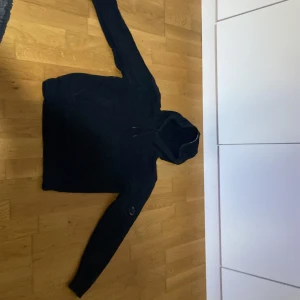 Svart hoodie från C.P. Company med en ficka på armen  - Säljer en stilren svart hoodie från C.P. Company. Den har en klassisk design med huva och långa ärmar. På ärmen finns en rund detalj med C.P. Company-loggan. Perfekt för en avslappnad look.