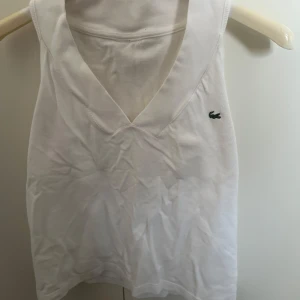 Vit ärmlös topp från Lacoste - Säljer en stilren vit ärmlös topp från Lacoste med v-ringning. Den har det klassiska gröna krokodilemblem på bröstet. Perfekt för en sportig look eller en varm sommardag.