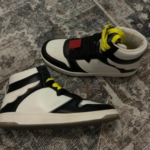 Svartvita sneakers med gula snören - Snygga svartvita sneakers med iögonfallande gula snören och en röd detalj på tungan. Skorna har en hög design med textdetalj på hälen. Perfekta för en trendig look. Aldrig använda!!!! 