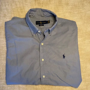 Ralph Lauren skjorta - Snygg blå skjorta från Ralph Lauren i slim fit. Skjortan har långa ärmar och knappar framtill. Den är tillverkad i bomull och har en broderad logotyp på bröstet.