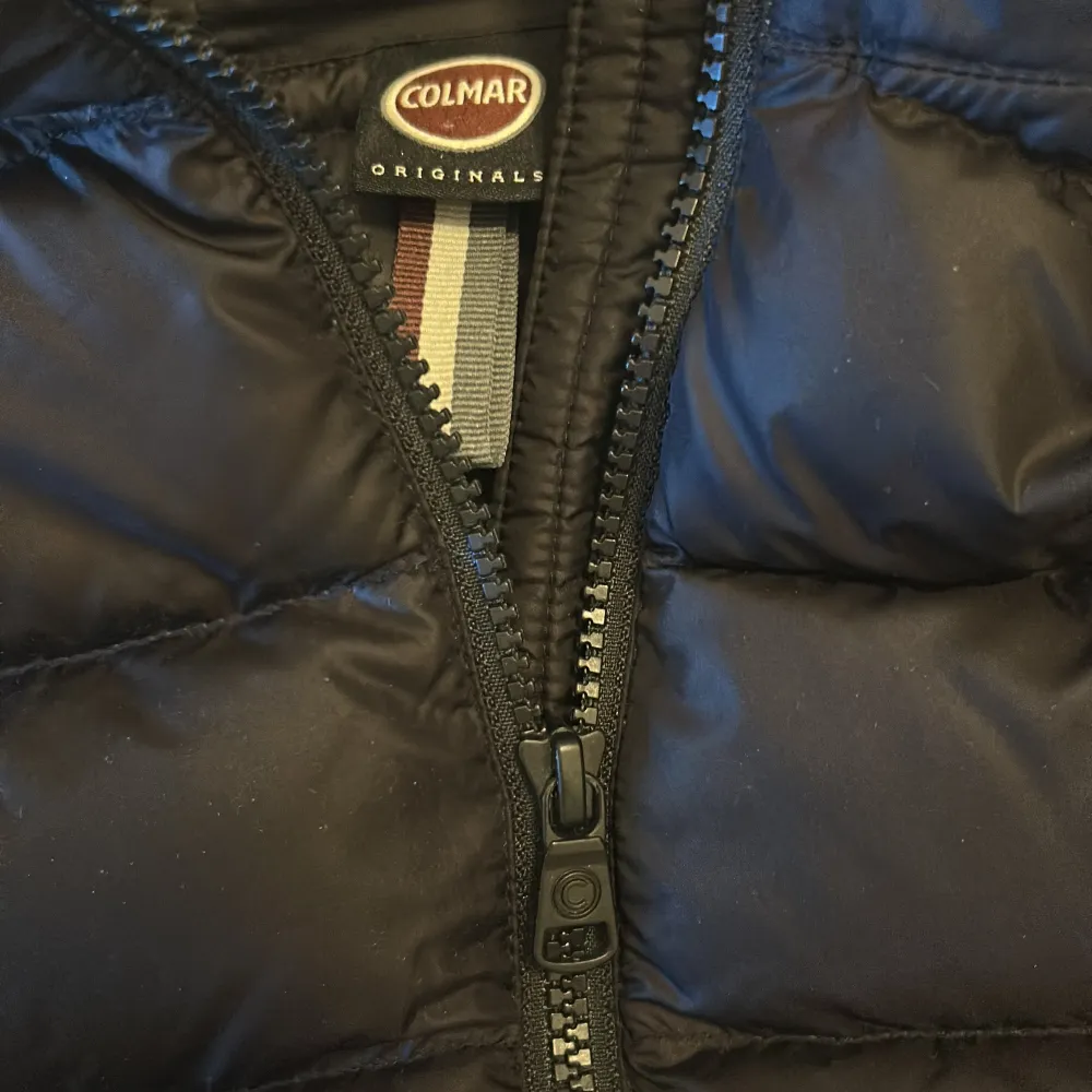 Snygg svart dunjacka från Colmar med huva och dragkedja. Jackan har en quiltad design och en rund logotyp på ärmen. Perfekt för kyliga dagar.. Takit.