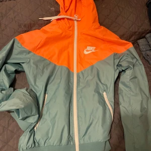 Grön och orange vindjacka från Nike - Säljer en snygg vindjacka från Nike i grönt och orange. Jackan har en dragkedja framtill och två praktiska fickor med dragkedjor. Perfekt för blåsiga dagar med sin lätta och bekväma design. Huvan har en justerbar dragsko för extra skydd.