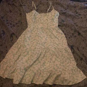 Blommig sommarklänning med volang - Charmig sommarklänning med blommigt mönster i ljusgrönt. Klänningen har smala axelband och volangdetaljer vid fållen. Perfekt för varma dagar!