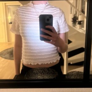 Vit ribbad topp - Säljer en vit ribbad topp med korta ärmar och en hög hals. Toppen har en elastisk struktur som ger en bekväm passform och en vågig kant vid ärmslut och nederkant.