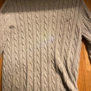 Beige kabelstickad tröja från GANT - Säljer en stilren grå kabelstickad tröja från GANT i premium bomull. Tröjan har en klassisk design med rund hals och långa ärmar. Perfekt för en avslappnad och elegant look. Obs! Priset är inte hugget i sten.