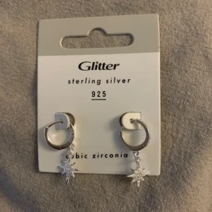 Silverörhängen med stjärndetalj från Glitter - Snygga silverörhängen från Glitter med cubic zirconia och en hängande stjärndetalj. Tillverkade i sterling silver 925, dessa örhängen ger en elegant touch till din stil.