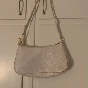 Vit axelväska med krokodilmönster - Snygg vit axelväska med krokodilmönster och gulddetaljer. Väskan har en praktisk dragkedja upptill och en justerbar axelrem. Perfekt för att ge en stilren touch till din outfit.